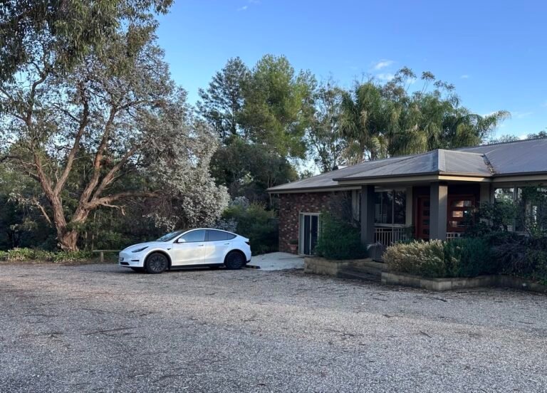 White Tesla Model Y parked on a gravel driveway in front of a suburban house with trees
