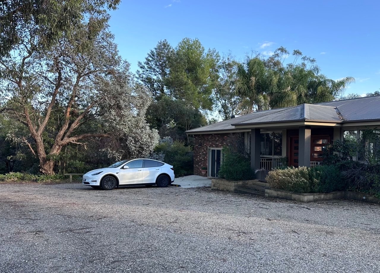 White Tesla Model Y parked on a gravel driveway in front of a suburban house with trees