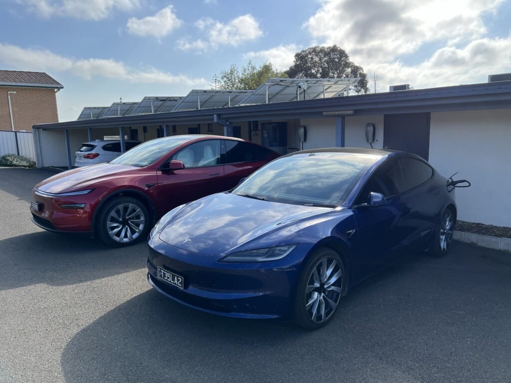 Red Tesla Model Y and blue Tesla Model 3 at motel wall chargers beneath rooftop solar panels